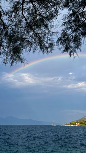 A vertical shot of a rainbow at the sea with a boat in distance.