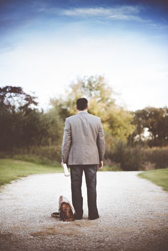 A male wearing a business suit standing on a pathway near his bag while holding the bible shot from behind