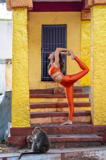 A vertical shot of a yogini doing Natarajasana in an ancient Indian temple with a monkey outside