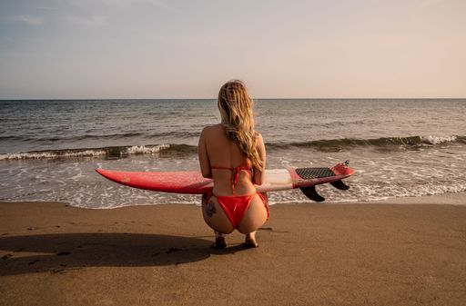 A Young woman with blonde hair walking on the beach with a surfboard on a sunny day