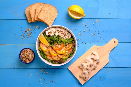 A top view of a bowl of mixed salad with tomatoes, mushrooms, lettuce, and seeds with bread on a wooden table