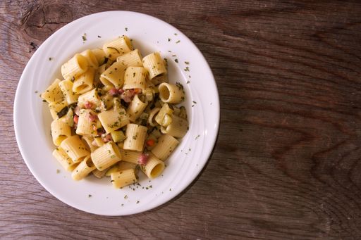 A closeup of rigatoni pasta with bacon and zucchini in a white bowl on the wooden background