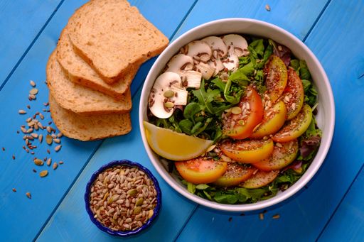 Top shot of a bowl of mixed salad with tomatoes, mushrooms, lettuce, and seeds with bread on a wooden table