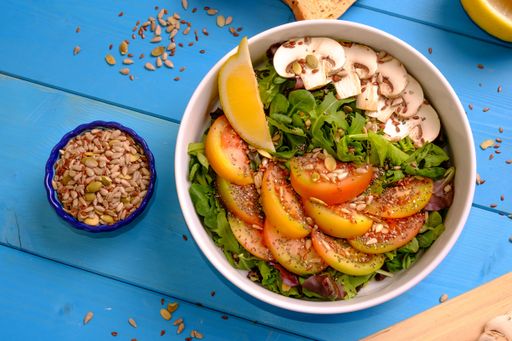A Top view of a bowl of mixed salad with tomatoes, mushrooms, lettuce, and seeds on a wooden table