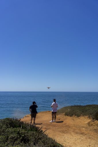 A closeup shot of two men flying a drone over a blue ocean standing on a cliff in Andalisia