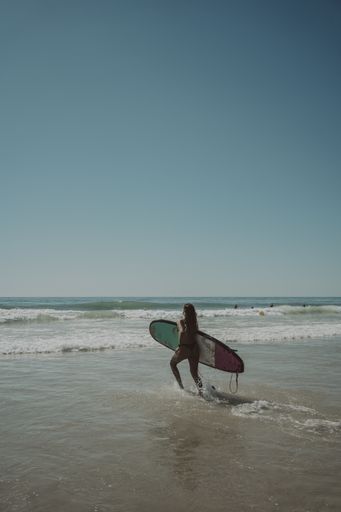 A vertical shot of a young lady running into the water with a surfing board