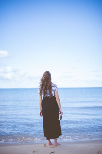 A vertical shot from behind of a female standing on a beach shore bare feet while holding the bible