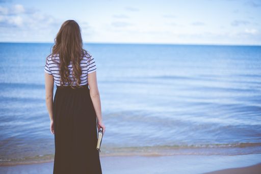 A female standing on the beach shore while holding the bible with a blurred background shot from behind