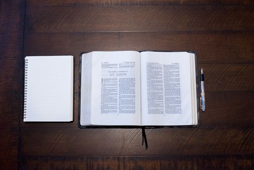 An overhead shot of an open bible in the middle of a notebook and a pen on a wooden surface