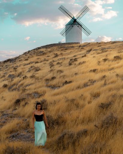 A vertical shot of a young brunette female walking in a dry grassland with a windmill