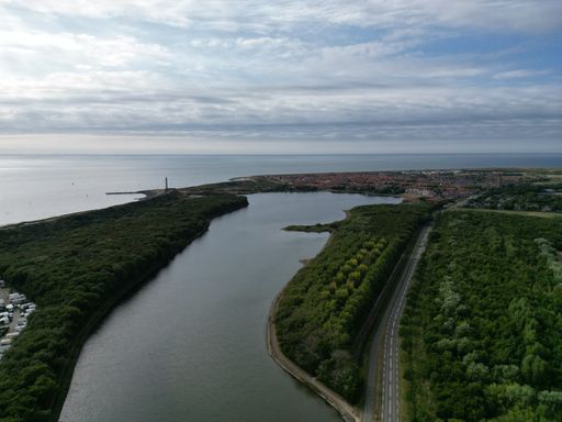 A drone shot over a river between green fields and cloudy sky on the horizon