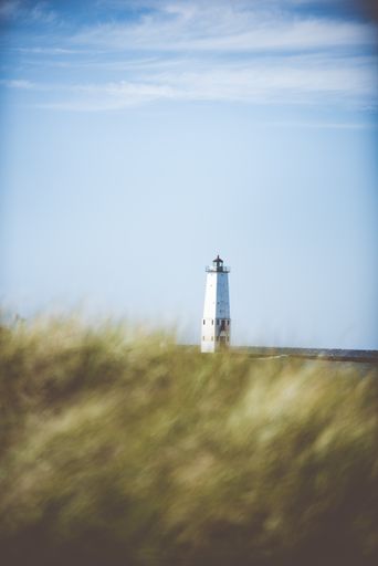 A vertical selective focus shot of a lighthouse in the distance with a blue sky in the background