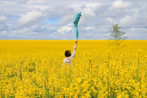 A female waving a blue scarf in the middle of a yellow field under the cloudy sky