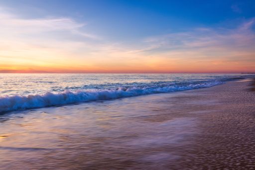 The sea waves splash over the shore at sunset with an orange cloudy sky in the background