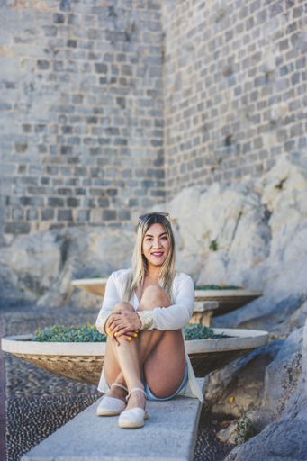 A young smiling Hispanic female sitting on a bench against an old wall under the sunlight