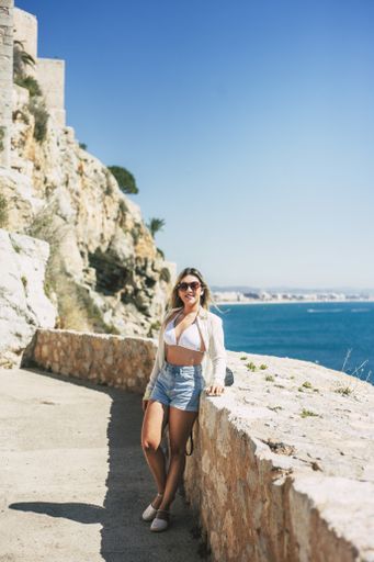 A vertical shot of a Hispanic female posing near the stone walls surrounded by the sea under the sunlight