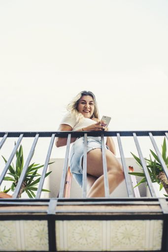 A low angle shot of a young Hispanic female using her phone and looking down from a balcony of a residential building