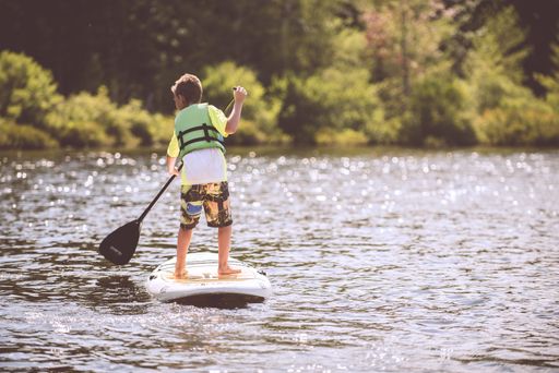 A child standing on a surfboard on the water while pedaling with a blurred background shot from behind