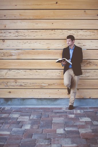 A vertical shot of a well-dressed male leaning against the wall while reading the bible