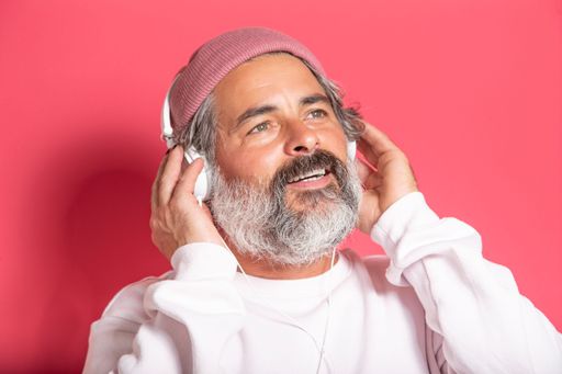 Grey-haired brunette man in sweater and casual hat listening to music with headphones isolated on pink background studio portrait