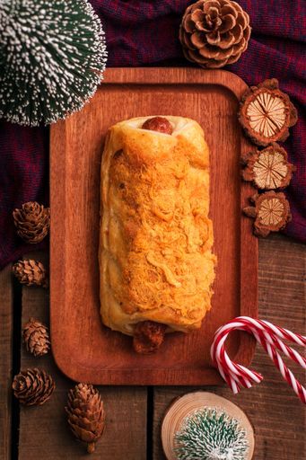 A top view of a baked sausage roll on a brown wooden board with Christmas decorations around