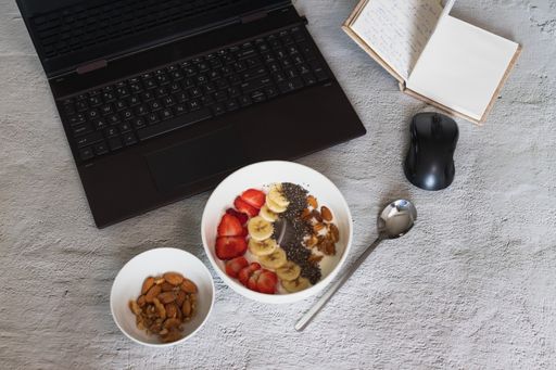 A high angle shot of a bowl of yogurt with fruits and nuts on the table with a laptop on it