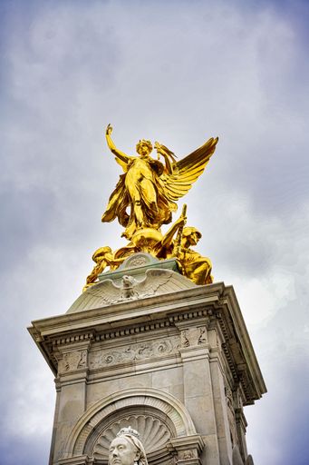 A low angle shot of Queen Victoria Memorial. Golden monument winged statue angel near Buckingham Palace, London, under cloudy sky