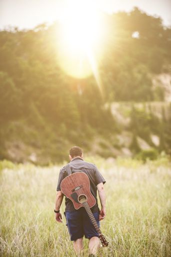A vertical shot of a male walking in a field next to a forest with a guitar on his back