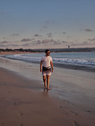 A vertical of a Caucasian man in a white shirt posing at the beach with the blue sea in the background