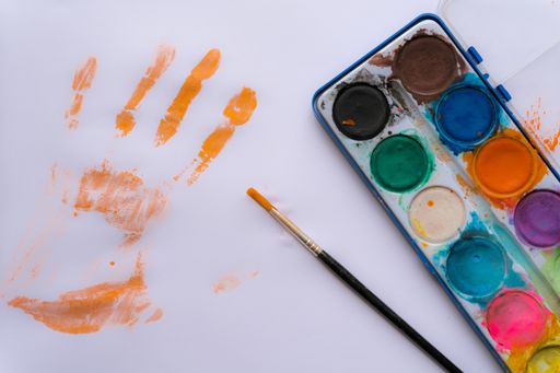 A top view of a watercolor palette, a brush and orange palm print on a white paper