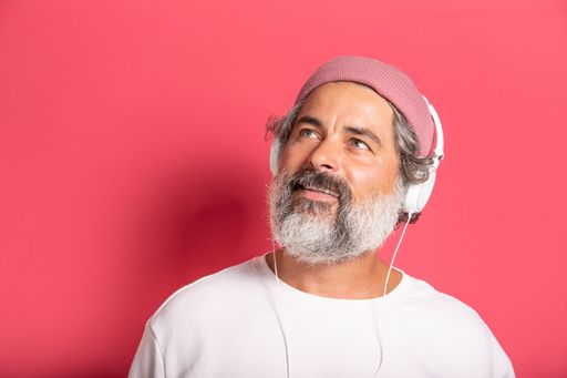 A Caucasian Cheerful gray-haired bearded man in casual pink hat and pullover listening to music with headphones isolated on fuchsia wall background