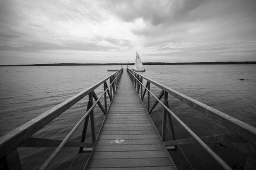 A grayscale shot of a wooden pier in the ocean with a single boat under the dark cloudy sky