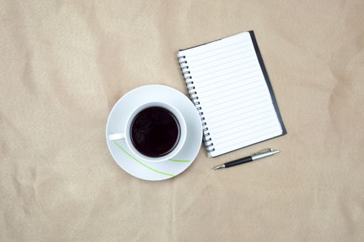 A top view of a cup of coffee on a wooden table with a pen and a notepad on it