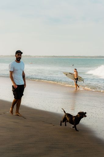 A vertical shot of a man with his dog on a  beach with a surfer and a sea in the background