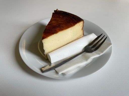 A piece of delicious cheesecake on a white plate with fork lying on a white table.