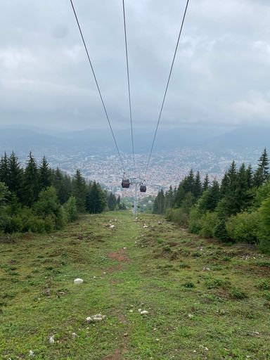 A vertical shot of the Sarajevo cable car over a green mount with trees and a city view in the background