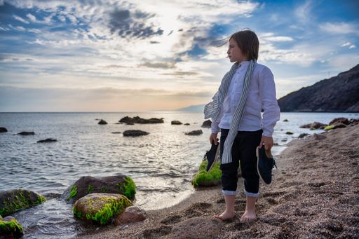 Boy poses on the beach with his shoes in his hands looking at the horizon.