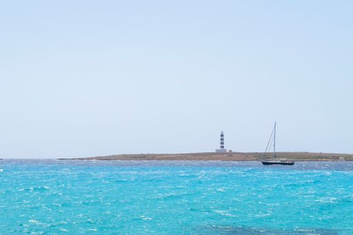 a sailboat skirts a cape with a blue and white lighthouse, on a sea of turquoise waters, Isla del Aire, Punta Prima, Menorca, Spain, copy space