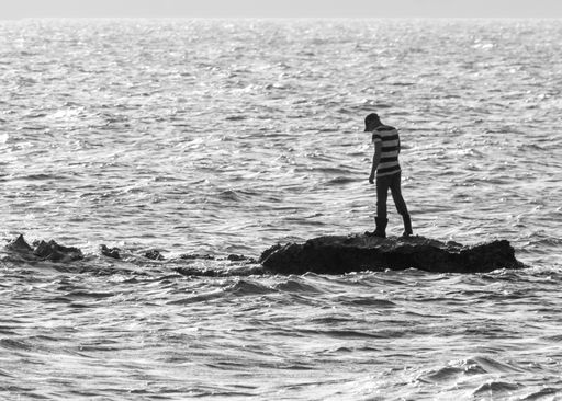 A grayscale shot of a male standing on a rock in the middle of the ocean