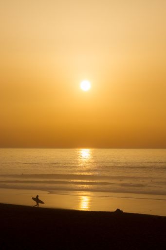 A vertical shot of a sunset over an ocean with a silhouette of a walking man with a board