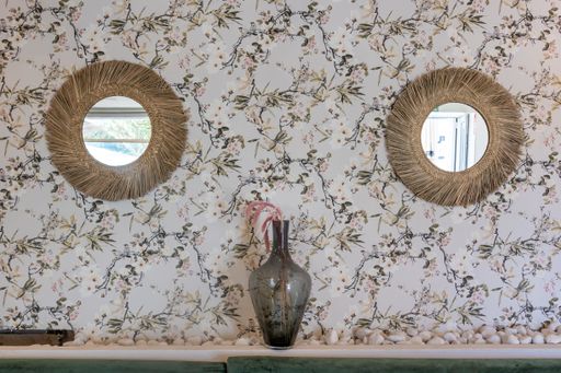 A vase put in the middle of two round mirrors hanging from a flower-printed wall