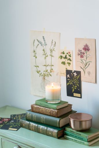 A vertical shot of a scented candle on top of stacked books with botanical posters on the wall