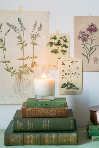 A vertical shot of a scented candle on top of stacked books with botanical posters on the wall