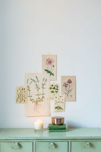 A vertical shot of a scented candle on top of stacked books with botanical posters on the wall