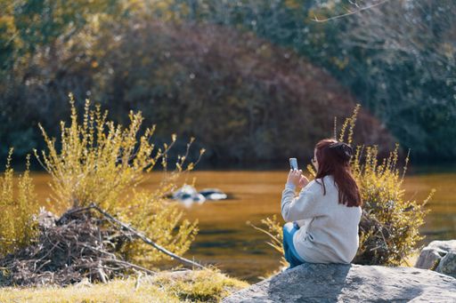 A Caucasian girl sitting by the lake and taking photo with her smartphone