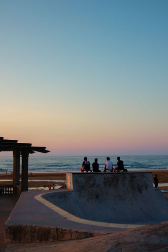 A back shot of young men sitting with their skateboards on the edge of a skate yard at the beach at sunset