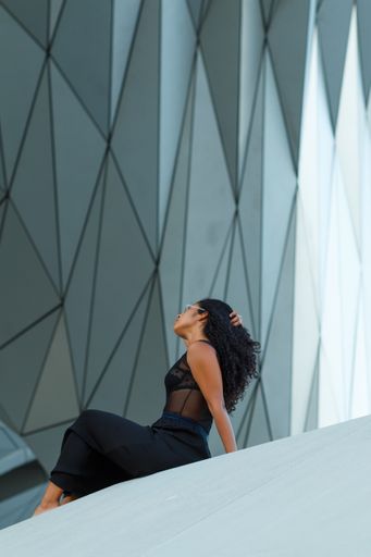 A vertical shot of a curly brunette Hispanic female an elegant black dress sitting under the wall of a modern building with geometric patterns