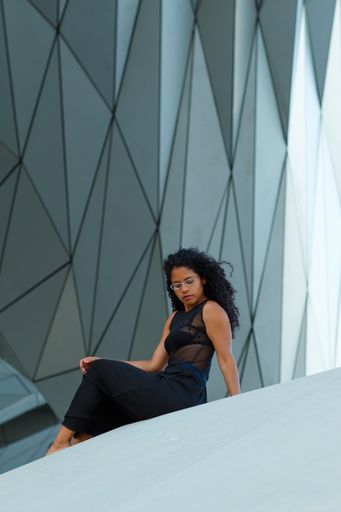 A vertical shot of a curly brunette Hispanic female an elegant black dress sitting under the wall of a modern building with geometric patterns