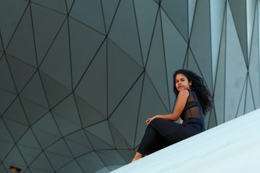 A curly brunette Hispanic female an elegant black dress sitting under the wall of a modern building with geometric patterns