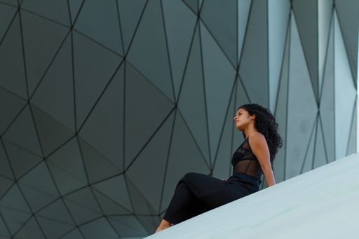A curly brunette Hispanic female an elegant black dress sitting under the wall of a modern building with geometric patterns